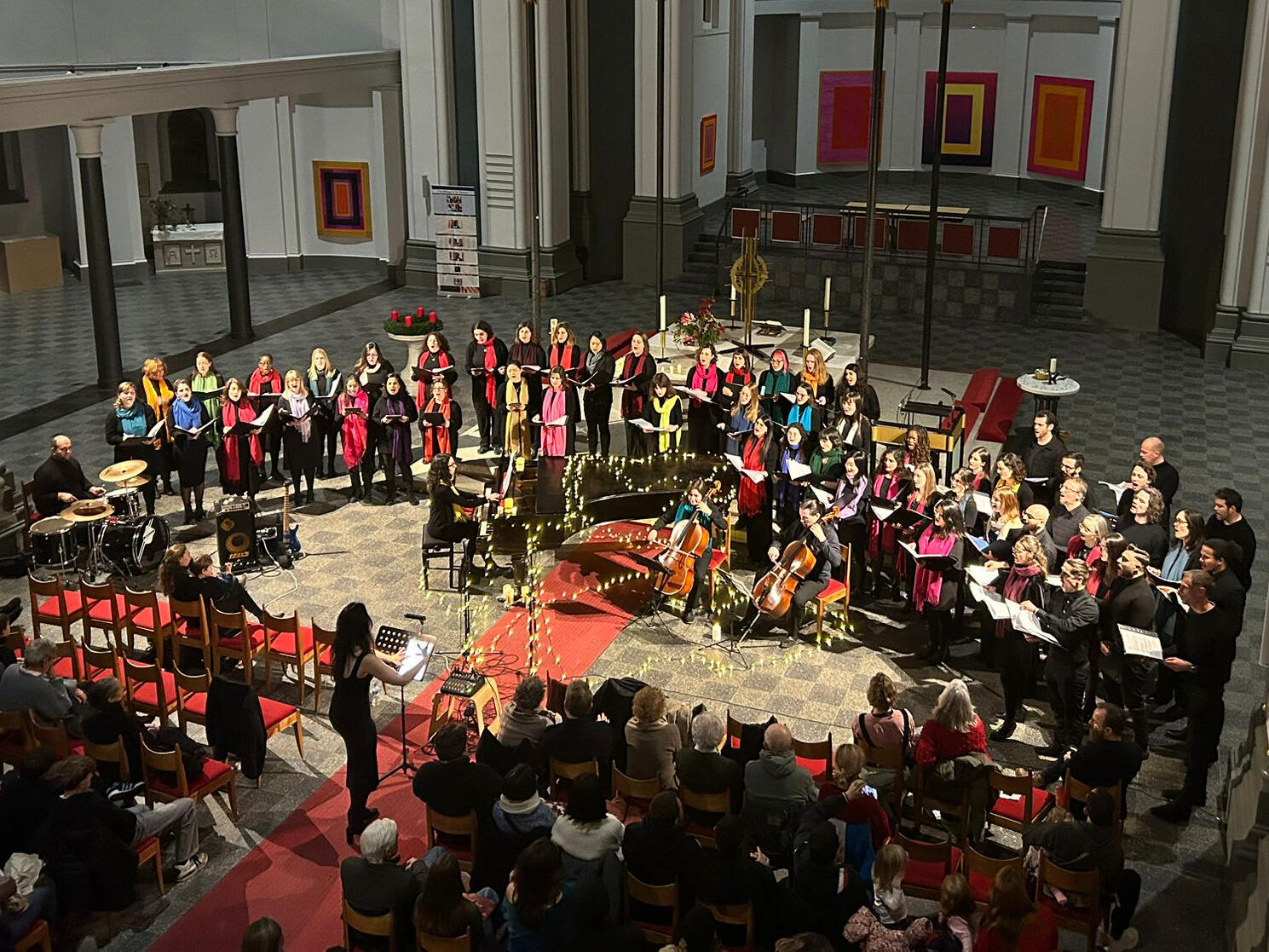 The full choir performs at their Christmas concert 2024 A choir with over 50 people dressed in black with colorful contrasting scarves perform in a half circle around a grand piano in a large church while the choir master conducts the choir.