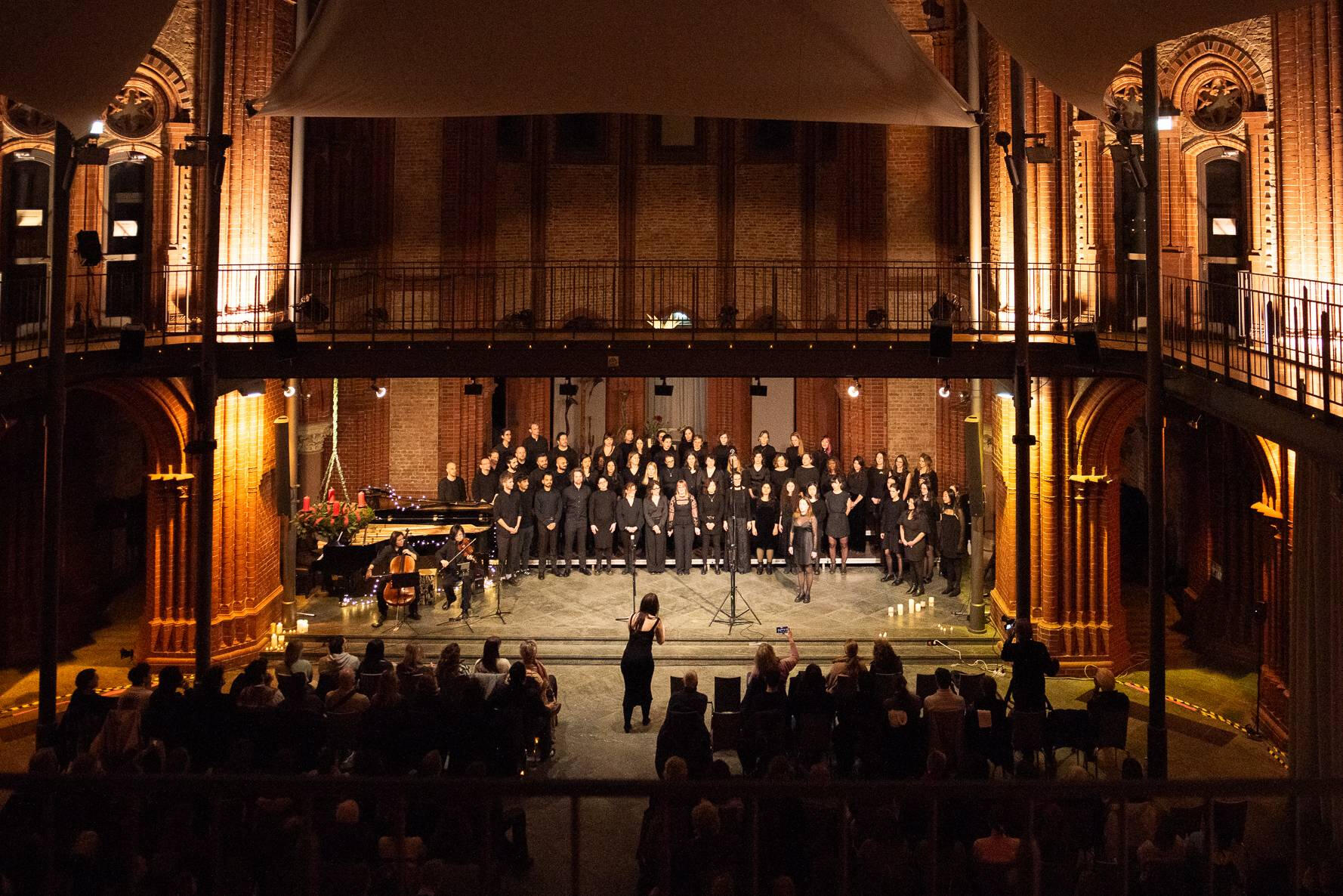The full choir performs at their Christmas concert 2025 A choir with over 60 people dressed in black perform in a large church while the choir master conducts. They are accompanied by a cellist and a violinist.