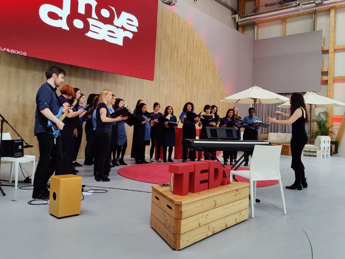 Part of the choir performing at TEDx Mitte in October 2024 Around 20 women dressed in blue tops and black bottoms perform on a stage with the TEDx logo in the foreground. They are accompanied by a man playing an electric guitar and the choirmaster conducting.